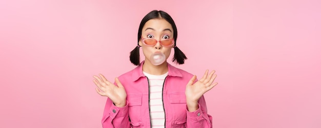 Stylish asian girl blowing bubblegum bubble chewing gum wearing sunglasses posing against pink background