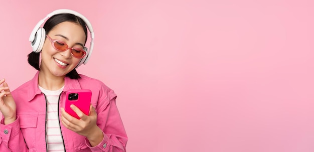 Free photo stylish asian girl dancing with smartphone listening music in headphones on mobile phone app smiling and laughing posing against pink background