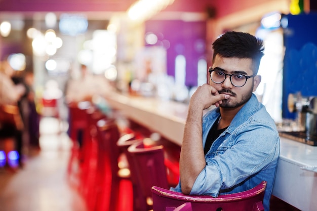 Stylish asian man wear on jeans jacket and glasses posed against bar in club.