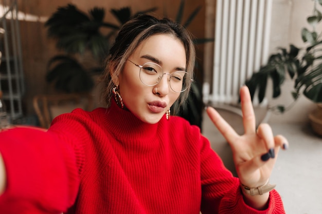 Free photo stylish asian woman in glasses gold earrings and red outfit winks takes selfie and shows peace sign