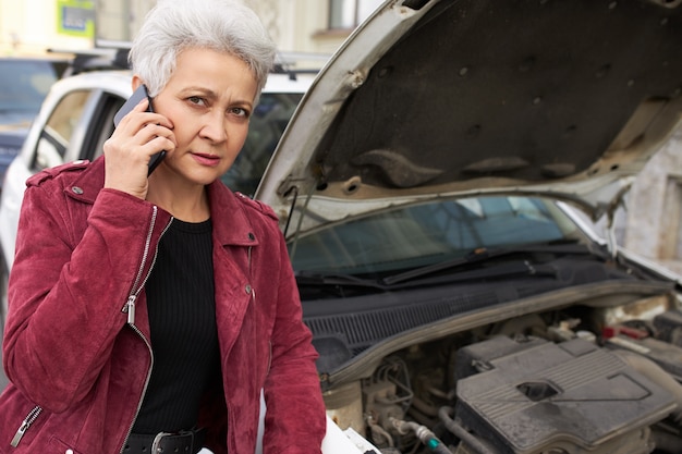 Free photo stylish attractive gray haired mature female driver standing near her broken white car with open hood and talking on the phone