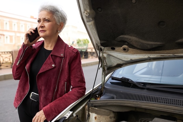 Free photo stylish attractive gray haired mature female driver standing near her broken white car with open hood and talking on the phone