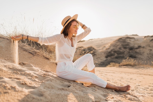 Free Photo stylish attractive smiling woman posing in desert sand dressed in white clothes outfit wearing straw hat and sunglasses on sunset