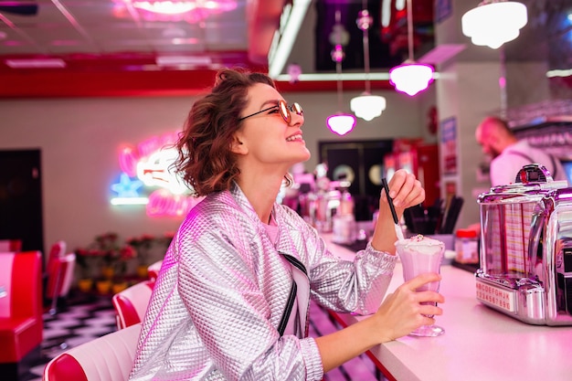 Free photo stylish attractive smiling woman in retro vintage 50's cafe of pink color sitting at table