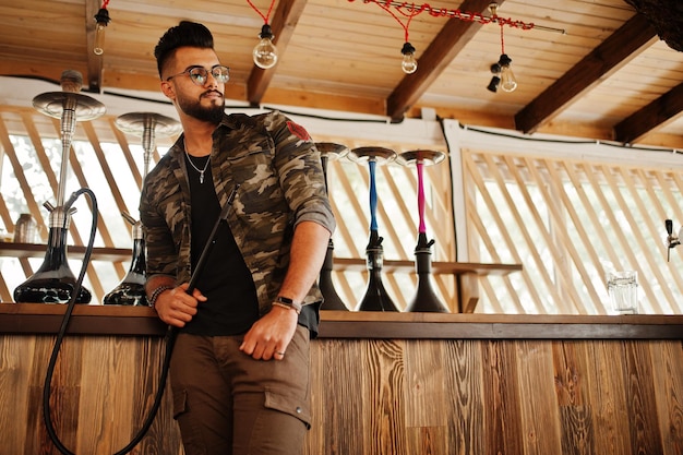 Stylish beard arabian man in glasses and military jacket smoking hookah at street bar Arab model having rest