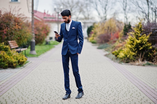 Stylish beard indian business man with bindi on forehead wear on blue suit posed outdoor looking at mobile phone