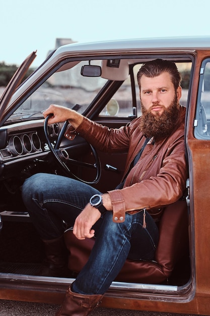 Free photo stylish bearded male dressed in brown leather jacket sits behind the wheel of a tuned retro car with open door.