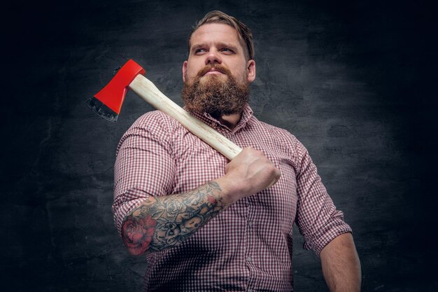 Stylish bearded male with tattoos on arms wearing pink plaid shirt and holds an axe.