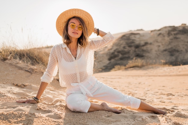 Stylish beautiful woman in desert sand in white outfit wearing straw hat on sunset