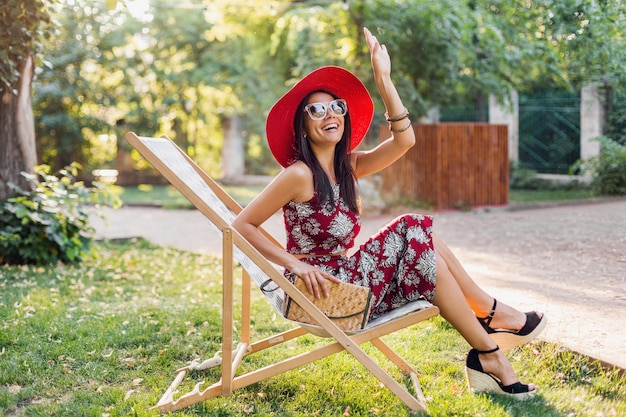 Stylish beautiful woman sitting in deck chair in tropical style outfit, waving hand, summer fashion trend, straw handbag, red hat, sunglasses, accessories, smiling, happy mood, vacation