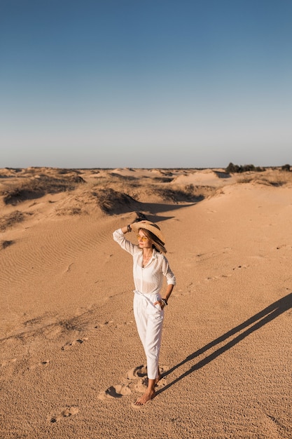 Free Photo stylish beautiful woman walking in desert sand in white outfit wearing straw hat on sunset