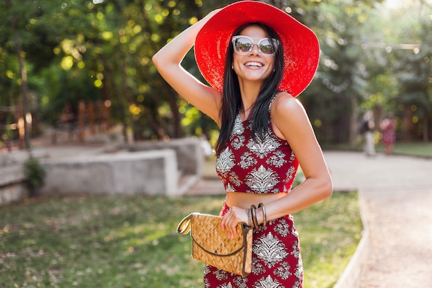 Stylish beautiful woman walking in park in tropical outfit. lady in street style summer fashion trend. wearing straw handbag, red hat, sunglasses, accessories. girl smiling in happy mood on vacation.