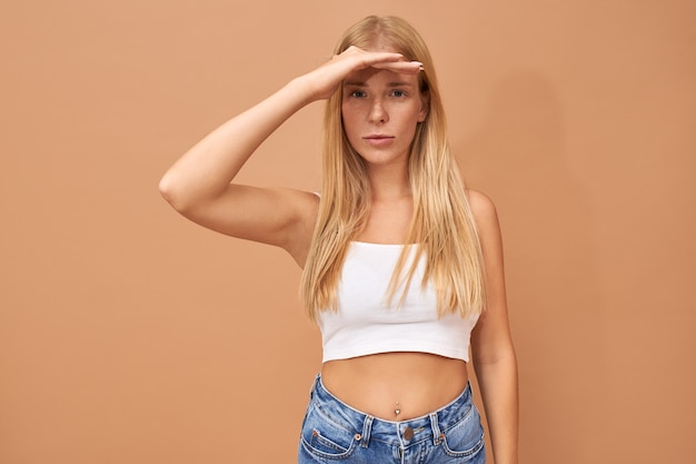 Free photo stylish beautiful young woman in white top and blue jeans keeping palm on her forehead, protecting eyes from bright sunshine