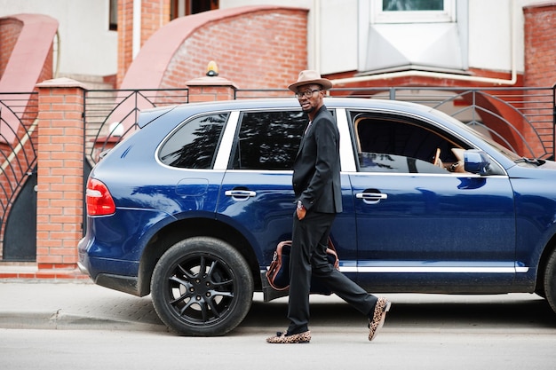 Free photo stylish black man at glasses with hat wear on suit with handbag against luxury car rich african american businessman