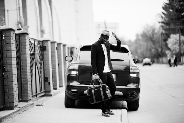 Stylish black man at glasses with hat wear on suit with handbag against luxury car Rich african american businessman