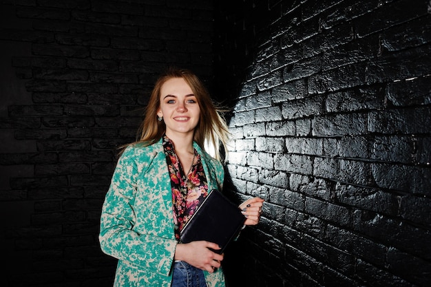 Stylish blonde girl in jacket and jeans with notebook diary at hands against brick black wall at studio