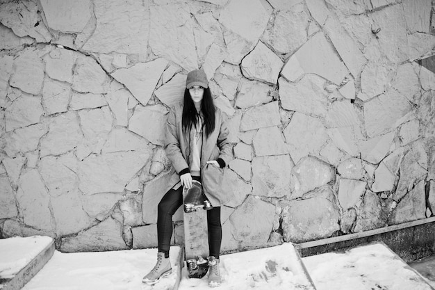 Stylish brunette girl in gray cap casual street style with skate board on winter day against colored wall