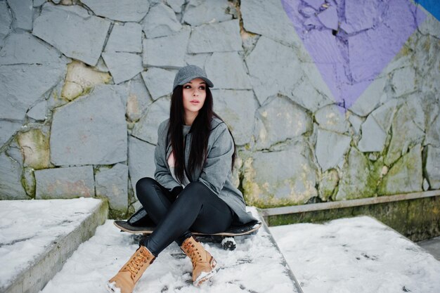 Stylish brunette girl in gray cap casual street style with skate board on winter day against colored wall