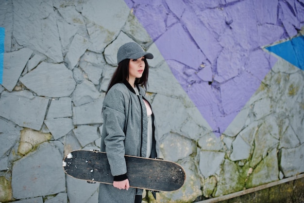 Free photo stylish brunette girl in gray cap casual street style with skate board on winter day against colored wall