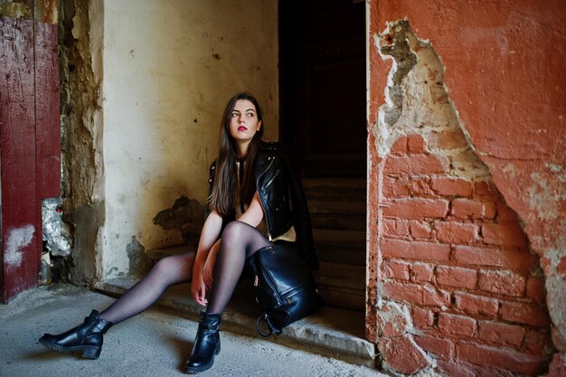 Stylish brunette girl wear on leather jacket and shorts with backpack sitting on old stairs against orange wall