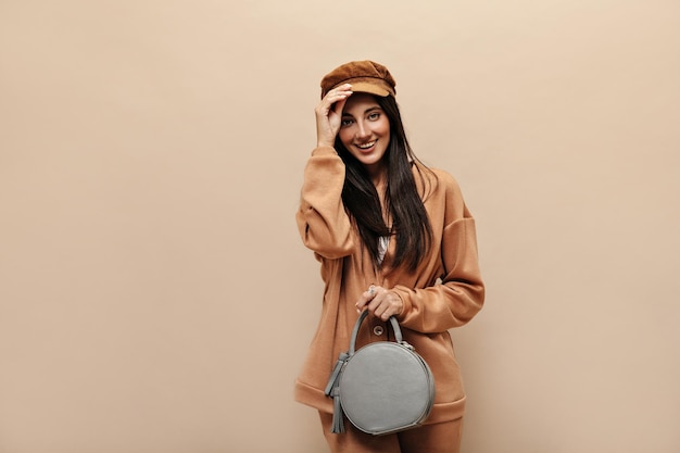 Free Photo stylish brunette longhaired woman in sport suit holds grey round handbag pretty girl in cap poses on isolated beige background