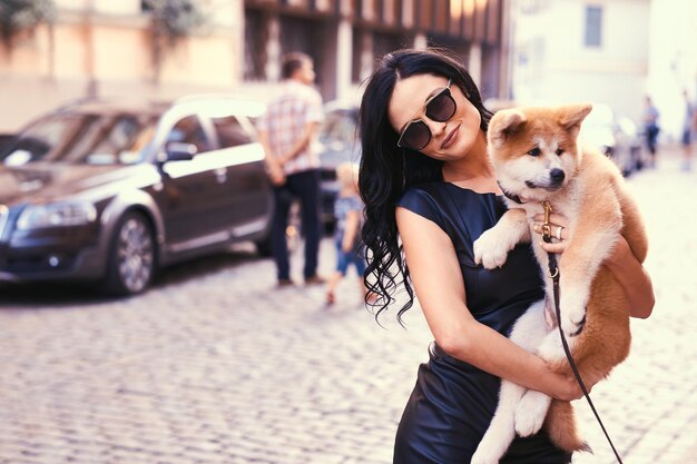 A stylish brunette woman in a black dress and sunglasses, standing on a street and holds a cute purebred puppy Akito.