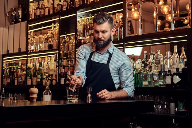 Stylish brutal bartender in a shirt and apron makes a cocktail at bar counter background.