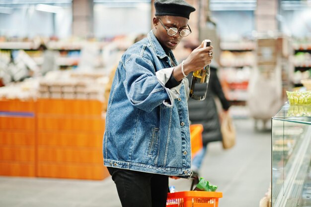 Stylish casual african american man at jeans jacket and black beret holding basket and looking on bottle of wine shopping at supermarket