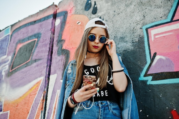 Free Photo stylish casual hipster girl in cap sunglasses and jeans wear listening music from headphones of mobile phone against large graffiti wall