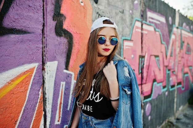 Free photo stylish casual hipster girl in cap sunglasses and jeans wear listening music from headphones of mobile phone against large graffiti wall