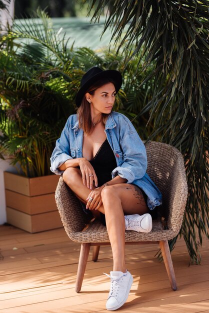 Stylish caucasian woman in dress, hat and denim jacket sit on chair