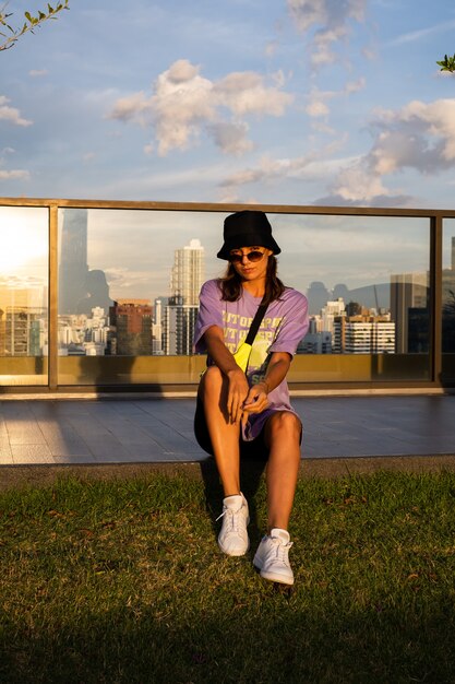 Stylish caucasian woman in trendy panama and waist neon bag on roof in Bangkok with view