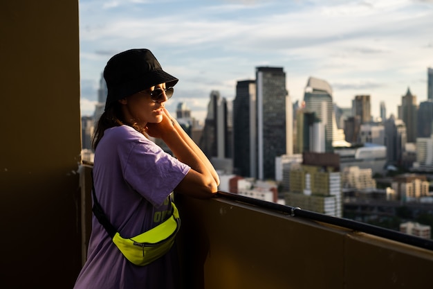 Stylish caucasian woman in trendy panama and waist neon bag on roof in Bangkok