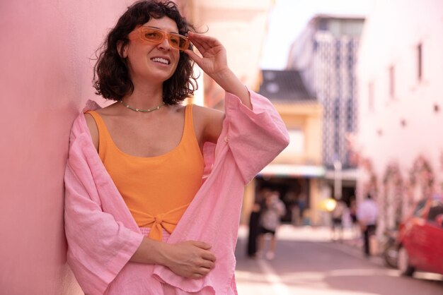 Stylish caucasian young brunette woman in pink summer clothes looks to side through glasses standing on city street