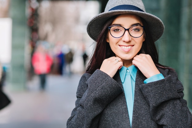 Stylish city portrait young pretty woman in grey hat, black glasses walking on street in centre. Luxury coat, fashionable model, cheerful emotions, smiling. 