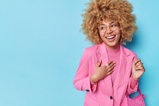 Free Photo stylish curly haired woman dressed in elegant pink jacket carries bag smiles broadly looks away glad to hear compliment isolated over blue background blank space for your advertising content