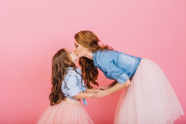 Free photo stylish curly mom and daughter hold hands and kiss sweetly at children's event on the pink background. little long-haired girl in denim shirt and lush skirt kissing her young mother at birthday party