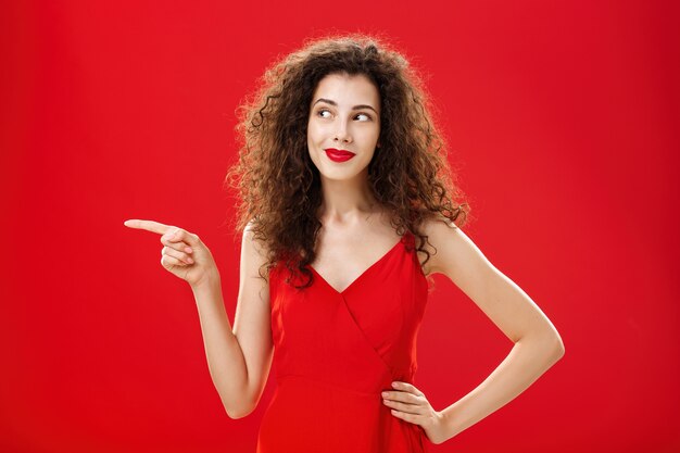 Stylish and elegant curious charming woman with curly hairstyle in red lipstick and trendy dress holding hand on hip pointing and looking left delighted and interested over studio background.