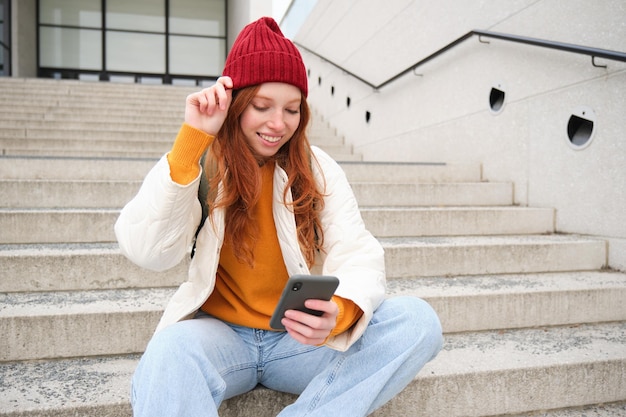 Stylish european girl with red hair sits on public stairs with smartphone places online order sends