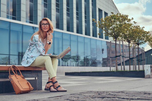 Stylish fashion blogger relaxing outdoor, working on the laptop, sitting on a bench against a skyscraper.