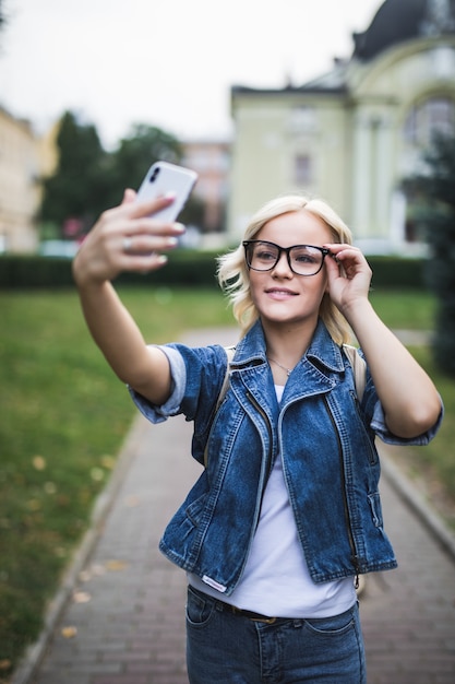 Stylish fashion blonde girl woman in jeans suite and glasses makes selfie on her phone in the city in the morning