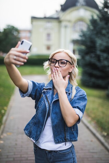 Stylish fashion blonde girl woman in jeans suite makes selfie give airkiss on her phone in the city in the morning