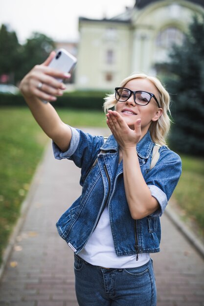 Stylish fashion blonde girl woman in jeans suite makes selfie give airkiss on her phone in the city in the morning