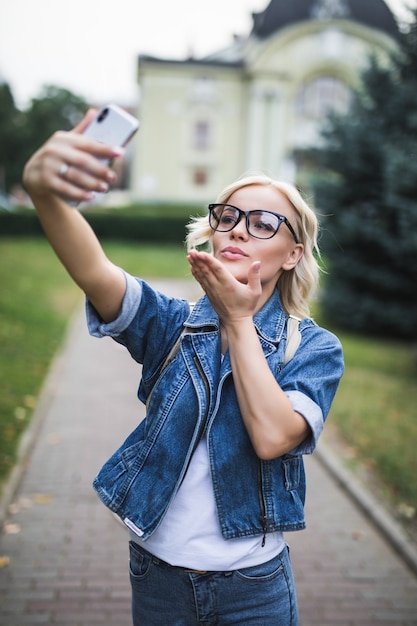 Stylish fashion blonde girl woman in jeans suite makes selfie give airkiss on her phone in the city in the morning