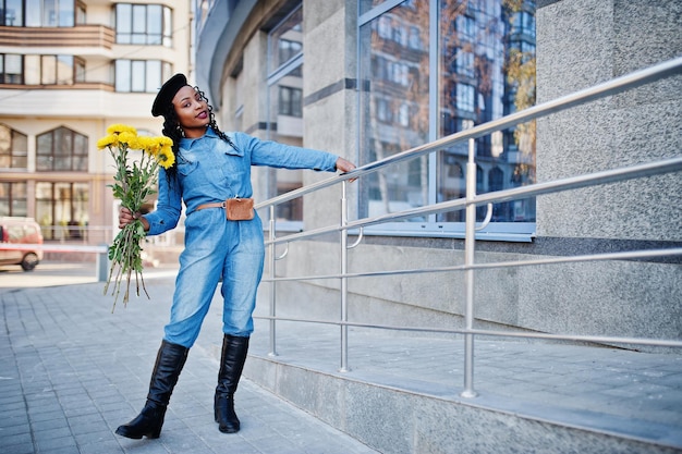 Free photo stylish fashionable african american women in jeans wear and black beret with yellow flowers bouquet posed outdoor in sunny day against blue modern building