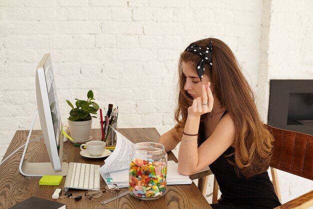 Stylish female student of school of economics working on diploma project, sitting at her workspace at home with computer, paper sheets and inteior items on table, eating sweets out of glass jar