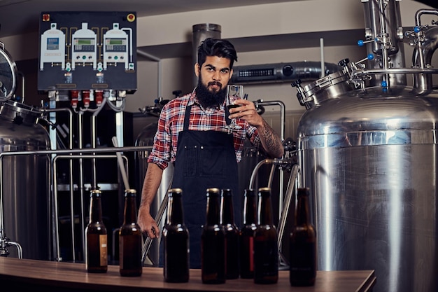 Free Photo stylish full bearded indian man in a fleece shirt and apron holds a glass of beer, standing behind the counter in a brewery.