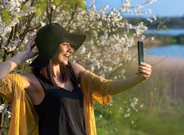 Free Photo a stylish girl in a hat makes a selfie at sunset near flowering trees in the forest