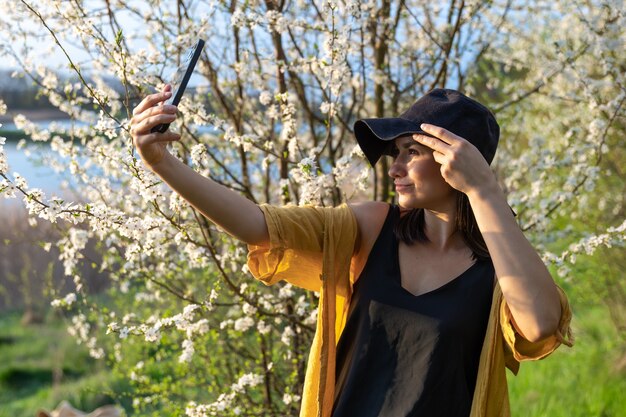 A stylish girl in a hat makes a selfie at sunset near flowering trees in the forest