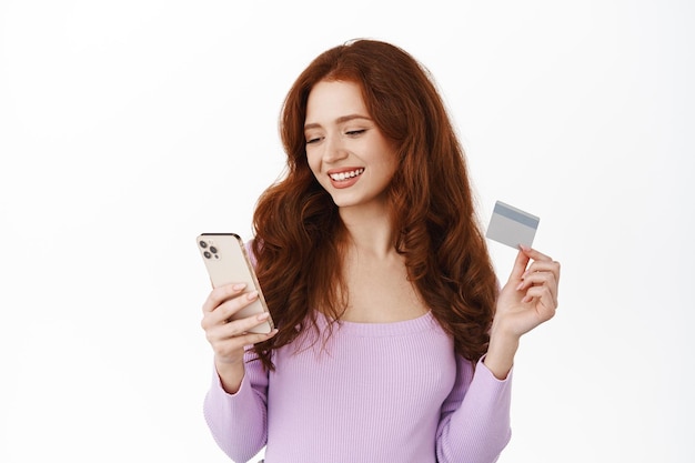 Free Photo stylish girl paying in mobile phone, order something, holding credit card with smartphone and shopping online, smiling relaxed, standing over white background